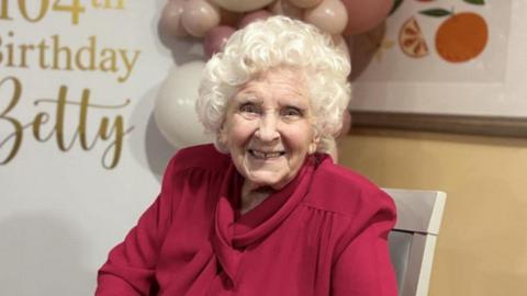 Elizabeth Milner sits in a white chair as she smiles at the camera. She has short curly white hair and wears a dark red blouse. 