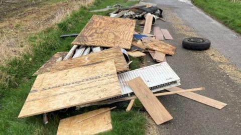 A large amount of building waste, including timber, a radiator, a vehicle tyre and wood dumped on the side of a country road.