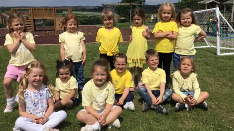 A group of young children on a school playing field wear yellow clothes or ribbons. They sit on the grass and pose for a photograph