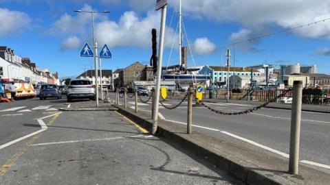 A photo which shows an area called The Bridge in Guernsey. There are metal poles which are acting as a fence in the middle of the photo, and two car parking spaces shown.