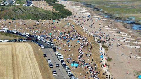 A large crowd is spread across a beach and grassy coastal area near Goring-by-Sea in West Sussex. Long lines of parked cars stretch along a road beside the shoreline while people gather to watch the eclipse.