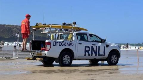 A white RNLI vehicle parked on the sand. There is a lifeguard, dressed in red, stood on the back looking out to the sea. There are people in the water. On the side of the vehicle it has the words 'Lifeguard' and 'RNLI'. There is a surfboard on the back of the vehicle.