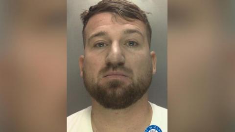 Dawid Dembler is pictured in a mugshot wearing a white t-shirt. He has light brown hair and a light brown beard.
