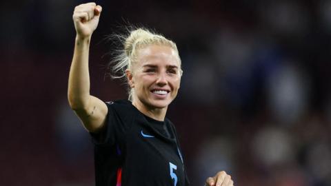 Alex Greenwood has blonde hair tied back in a ponytail. She has one arm raised in the air and is smiling at the crowd