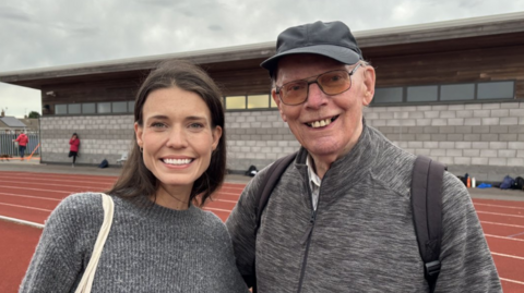 The photo shows a woman and a man smiling at the camera. The woman, on the left, has brown long hair and is wearing a grey jumper. The man, on the right, has a grey jacket on. He's also wearing glasses and a grey cap. They're standing on a brown athletics track. There is also a grey concrete building behind them.