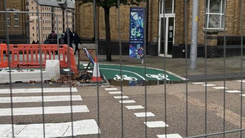 A view through a fence of where the cycle path meets the road at a zebra crossing. The end of the cycle path is flooded.