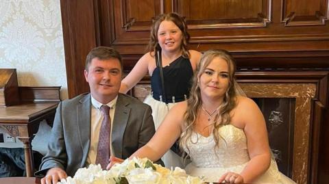 Wedding photo of Derek and Kylie Smith. Derek is clean-shaven and wears a grey suit, white shirt and burgundy tie; Kylie has long blonde hair and wears a white strapless wedding dress and pendant necklace. The couple are holding hands, resting them on a table behind a bouquet of white flowers. Derek's daughter stands in the middle, behind the couple. She has long brown hair and wears a black and white dress. They are all smiling.