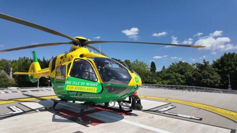A yellow and green helicopter sitting on a helipad surrounded by the tops of trees