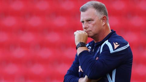 Stoke City manager Mark Robins watches from the touchline in his club sweatshirt. He has his left hand in a fist which is touching his chin.