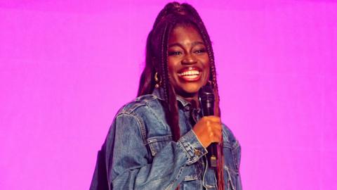Clara Amfo in a denim jacket smiling and holding a microphone to her face against a bright pink background.