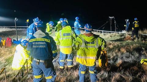 Coastguard crews gathered at the top of the cliffs at Marsden Grotto. There are 10 people dressed in helmets and hi-vis jackets. Floodlights have been set up in the grass and a rope has been deployed at the top of the cliff.