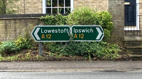 Green road direction signs. One points to the left and says Lowestoft A12 and the other points to the right and says Ipswich A12.