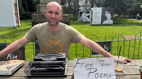 Patrick, a man with a shaved head and stubbled facial hair, smiles at the camera. He is sat on a wooden bench with a typewriter and a sign reading 'free typewriter poems' and is sitting in front of a patch of green.