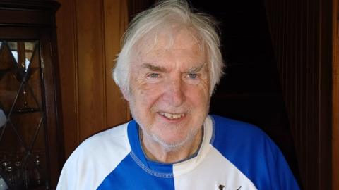 Jim Waters wearing a blue and white sports t-shirt. He has grey hair, a short grey beard and is smiling