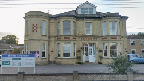 An Edwardian-looking yellow-brick large detached building. It has bow windows and a mansard window in the roof. In front of it is a car park and a low wall and a sign on the left saying The Dell Care Home. 