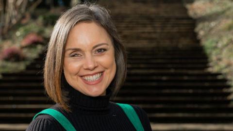 A woman with brown and grey hair sitting on some outdoor steps smiling. She has one eye and is wearing a black top with green overalls