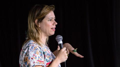 Lou Sanders wearing a colourful top and holding a microphone