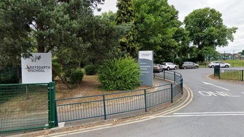 Main gate at Baysgarth School there are two signs with the school's name surrounded by green fencing and trees. A one way driveway is on the right with a number of parked cars