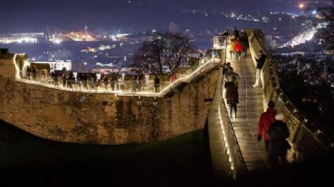 Lincoln Castle walls illuminated with warm lighting. It is evening and several people are walking around with coats, hats and scarves on. You can see the city lit up in the background.