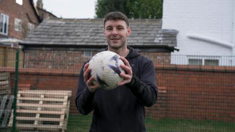 Man holding football looking at the camera with a wall and fence behind him.