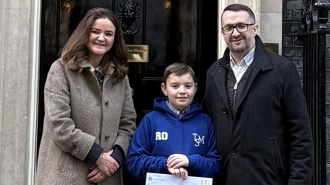 Roman stands in the middle of his dad Ryan and MP Jenny Riddell-Carpenter outside 10 Downing Street. Roman wears a blue hoodie, has short dark hair and holds a letter written on a piece of paper. Ryan has short dark hair, glasses and wears a black coat. Jenny has long dark hair and wears a long grey coat.