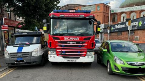 A fire engine, driving between a white Ford van and a green Vauxhall car, on a road in Luton.