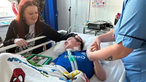 Woman with hand of head of teenage son in hospital bed, with nurse holding his hand