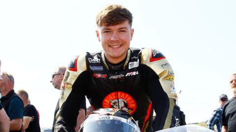 Storm Stacey sitting on his motorbike at a race weekend. He is smiling and wearing a black motorbike suit. 