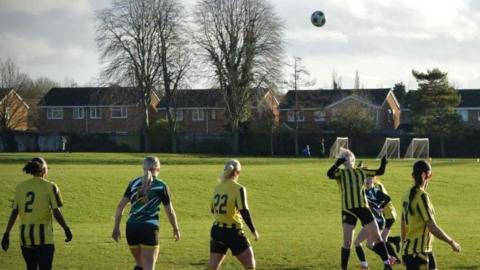 Girls in green shirts and yellow and black striped shorts play football on a football pitch. The football is high in the air, and one girl is jumping in the air mid-kick. Behind is the goal, and behind that are houses behind the field.