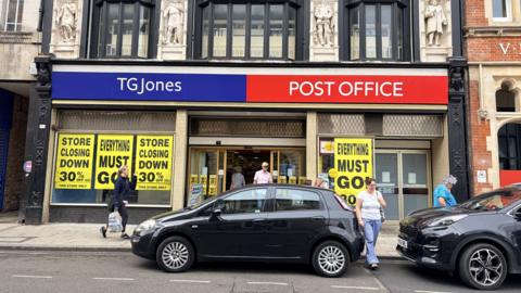 The front of a Post Office and TG Jones shop on a High Street. Cars are parked outside on the road and people are walking past as well as in and out of the shop.