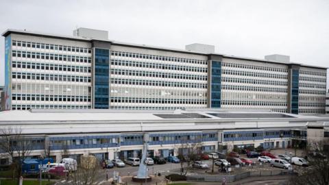 The outside of a large hospital, which is tall, blue and white with many windows, and car parked in front of it.