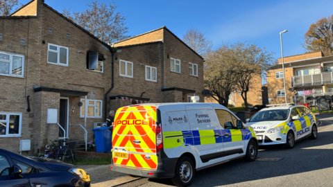Two police cars - including a forensics van - parked outside a block of terraced houses in Ipswich. A window of one of the homes is open and is smoke damaged.