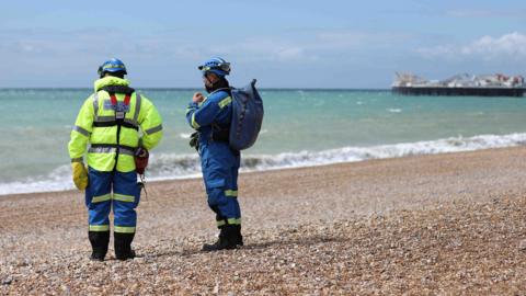 Two coastguard officers in yellow and blue on stoney beach with Brighton Pier in background