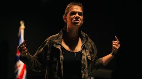 An actor in a black top and brown jacket stands on a dark stage with a Union flag in the background, gesturing and pulling an angry face, in the new play At Last.