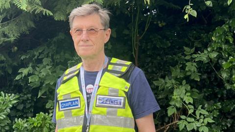 A man with grey hair and glasses looks off camera. He is wearing a blue T-shirt and a hi-vis waistcoat. On his right chest a blue badge says "CHAPLAIN". On the left chest a badge says "BRITISH TRANSPORT POLICE".