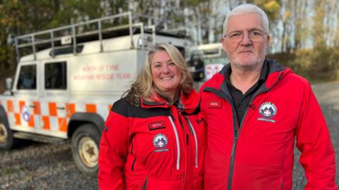 Volunteers Rachel Smith and Mark Silmon, wearing red mountain rescue uniform overalls, stand in front of a 4x4 rescue vehicle. Both of them are smiling looking into the camera. 