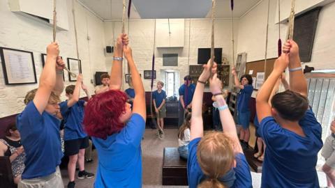 A group of young people are standing in a circle in a bell ringing tower. They are holding onto the ropes which are hanging from the ceiling above their heads. Most of the group are wearing blue T-shirts.