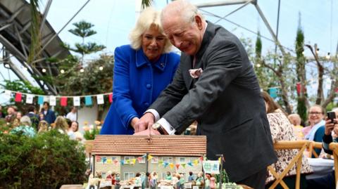 The King and Queen struggling to cut a cake together in the shape of terraced houses at the Eden Project. The Queen is wearing a blue dress, the king is in a grey suit. There are people eating lunch behind them, watching and smiling, and bunting hangs around the tables.