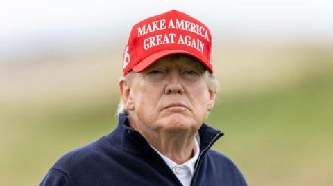 Head and shoulders of Donald Trump, who is wearing a white polo shirt, a navy blue fleece and a red cap which says "Make America great again"