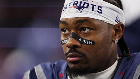Stefon Diggs of the New England Patriots watches his team play the New York Jets in November 2025. He wears his football uniform and athletic tape across his nose, on which he has written "Philippians 4:13," a reference to a Bible verse.