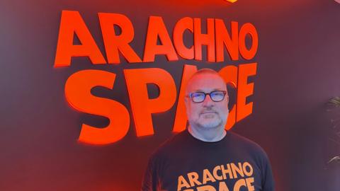 A man wearing glasses and a dark T-shirt is standing in front of a bright orange glow-in-the-dark sign that reads "Arachnospace".