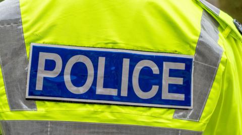 The back of a bright yellow high‑visibility police jacket with reflective strips and a blue patch displaying the word “POLICE” in bold white letters.