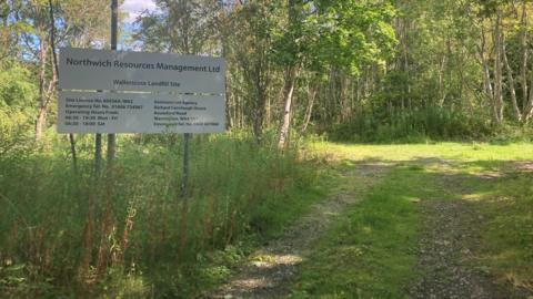 View of a sign for Wallerscote Landfill site. It has Northwich Resources Management written on the top, then Wallerscote Landfill Site beneath. There are also phone numbers and contact details on the site, which stands in overgrowth at the side of a gravel path.