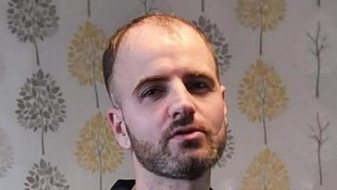Adam Lawson is balding and has a beard. He is wearing a black T-shirt and is looking at the camera. He is standing in front of wallpaper with grey and gold trees on it.