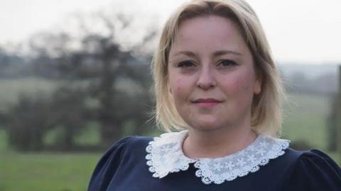 A woman with short blonde hair looking at the camera. She is stood in a field, which is blurred behind her. She is wearing a dark blue top with a white collar