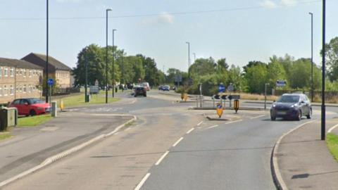 A road leading to a roundabout. Cars in the distance, trees on either side of the road. Houses on the left hand side of the road.
