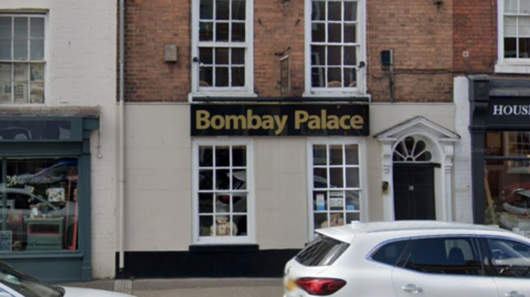 The exterior of Bombay Palace. It is a white building with a black and gold sign. Outside are two parked white cars with other shops either side.