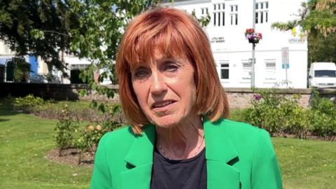 Woman with red hair, black top and green jacket standing in front of a park and a white building.