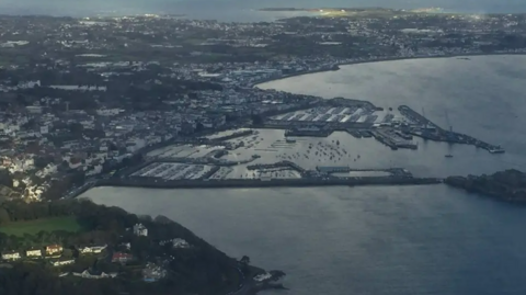 An aerial shot of Guernsey. There are houses on land on the left and the sea on the right.  