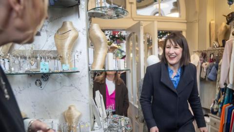 Chancellor Rachel Reeves walking into a shop in Leeds Corn Exchange, with a display of jewellery to the left. She is smiling and wearing a dark coat and a blue shirt. 